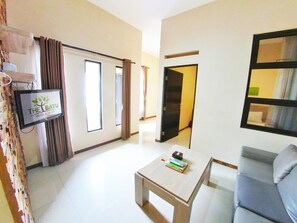 Een hotelkamer met een flatscreen-tv waarop het bord 'The Batu Hotel & Villas' staat, een bank, een salontafel en een deur die naar een andere kamer leidt.