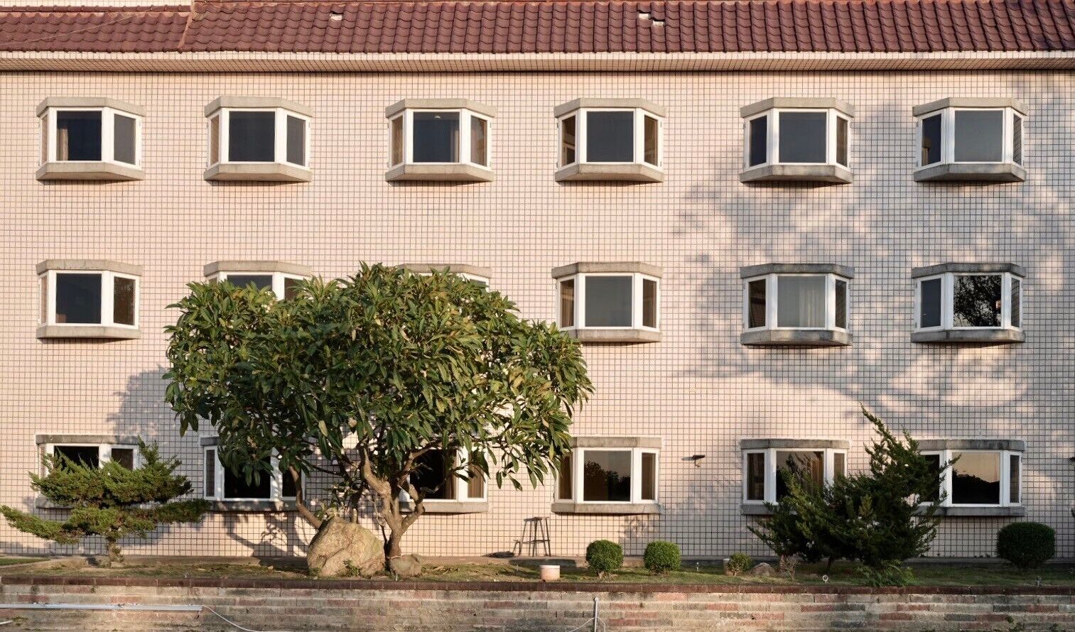 Un edificio de varios pisos con fachada de tejas, múltiples ventanas y un árbol en primer plano.