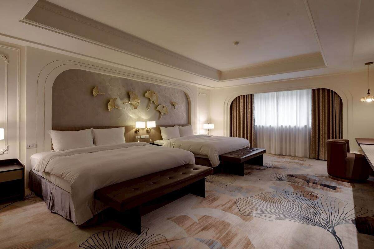 A hotel room with two beds, a bench, a desk, and a large window with curtains.