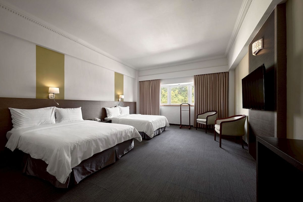 A hotel room with two beds, a desk, a chair, a television, and a window with curtains.