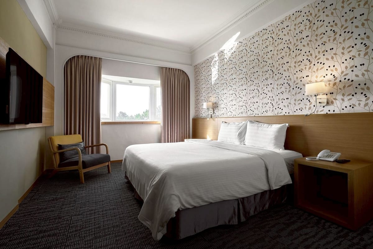 A hotel room with a bed, a chair, a nightstand with a phone, a window with curtains, and a wall with a leaf pattern.