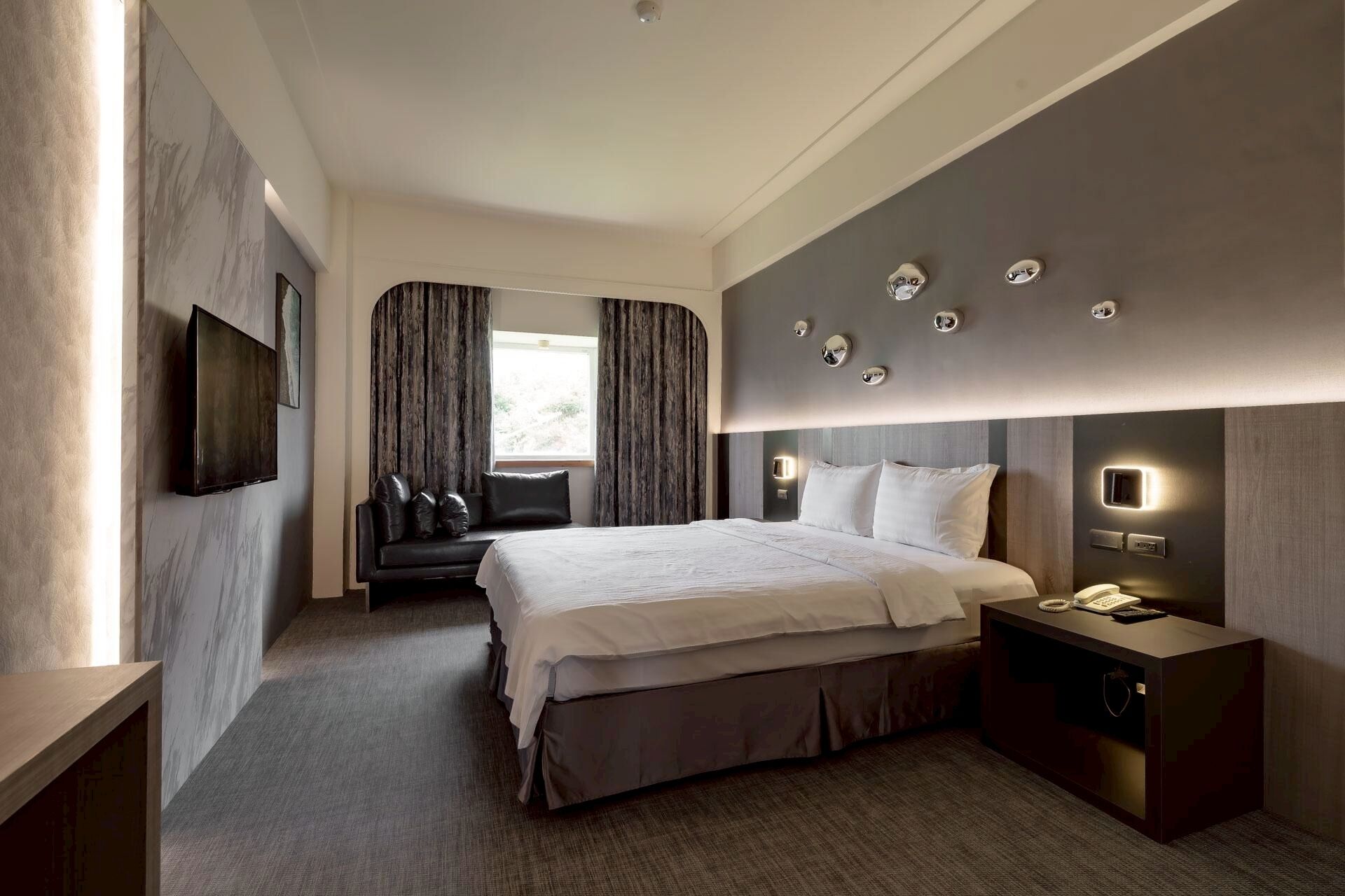 Quarto de hotel moderno com uma cama grande, um sofá, uma televisão e uma mesa de cabeceira com telefone.