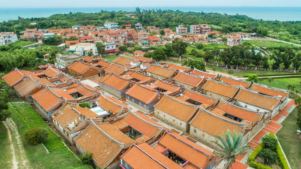 Un pueblo tradicional con edificios de tejados de terracota dispuestos en un patrón cuadriculado, rodeado de vegetación y vistas al mar.