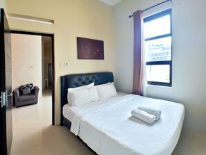 A hotel room with a large bed, a headboard, a window with curtains, a small seating area, and a painting on the wall.