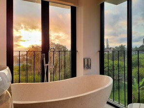 Un baño moderno con una bañera independiente, ventanas amplias y vistas a un jardín exuberante y edificios a la distancia.