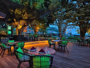 An outdoor dining area with round tables and chairs, surrounded by trees and illuminated by string lights. - The Batu Villas No 2 near Jatim Park (Batu)