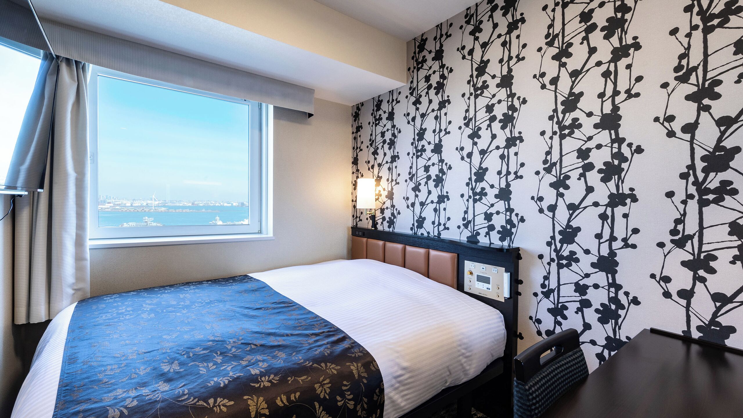 Una habitación de hotel con cama, ventana con vistas a la ciudad, pared con estampado floral y mesita de noche con lámpara.