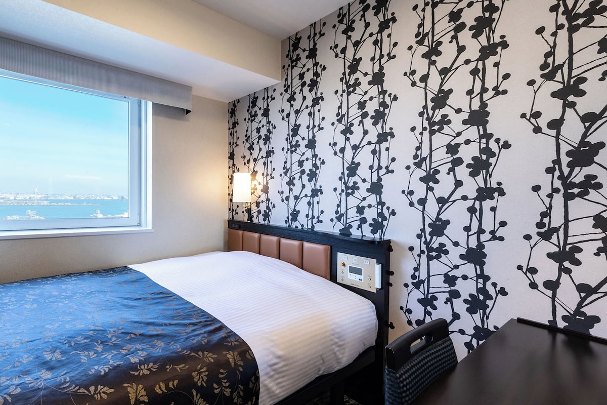 Ein Hotelzimmer mit Bett, Fenster mit Blick auf eine Stadtlandschaft, Wand mit Blumenmuster und Nachttisch mit Lampe.
