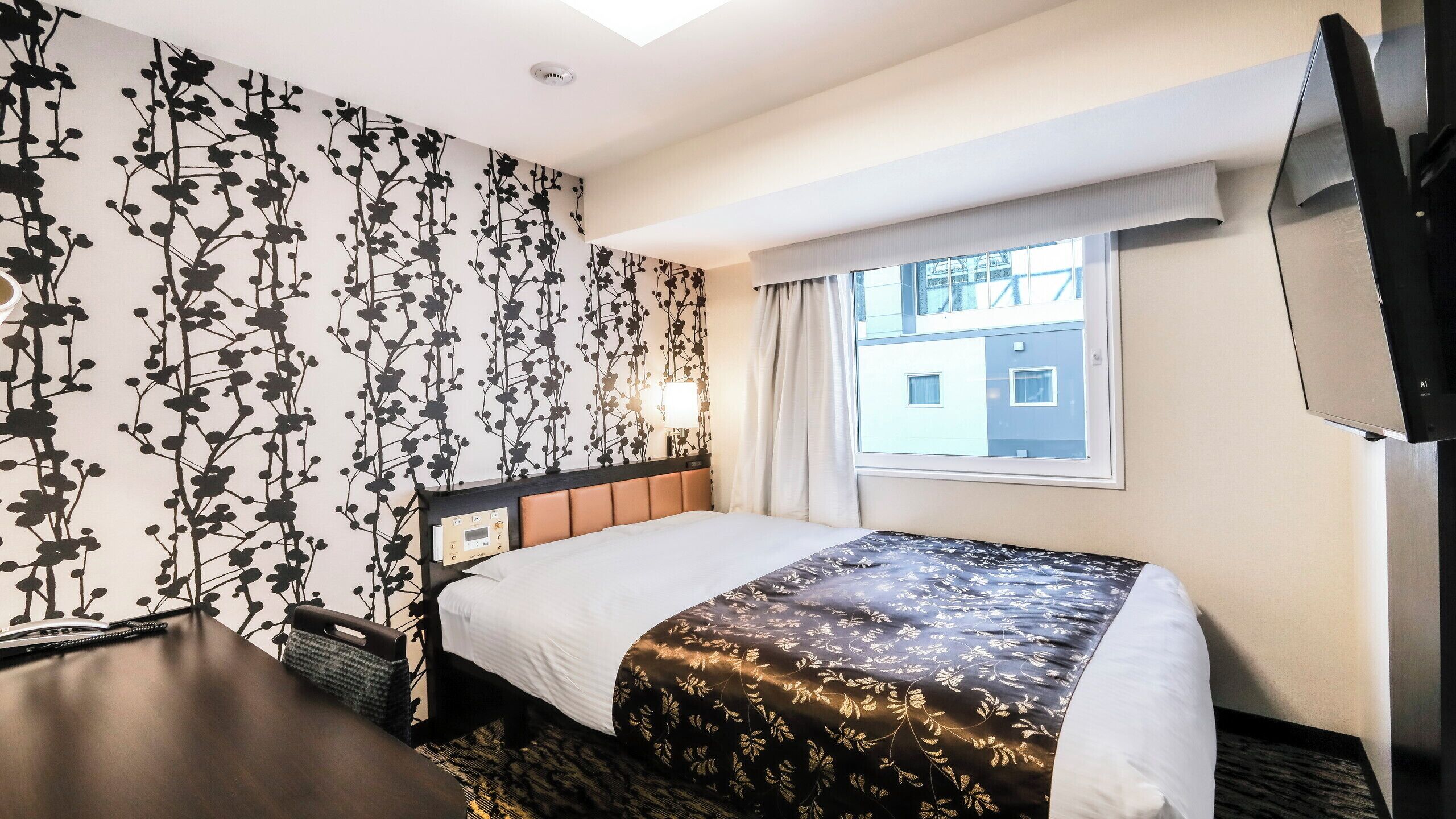 Quarto de hotel com cama, escrivaninha, cadeira, televisão e janela com vista para edifícios.