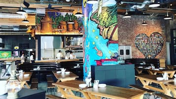 Un restaurante con mesas y bancos de madera, una cocina al fondo y un mural en la pared.
