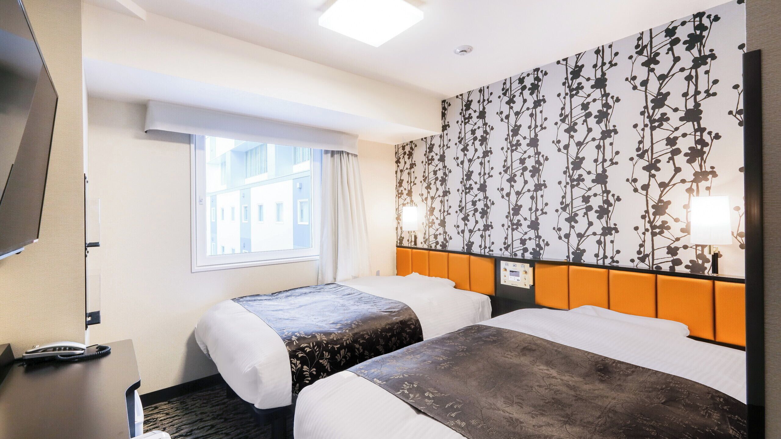 Quarto de hotel com duas camas, televisão, secretária com telefone e janela com cortinas.