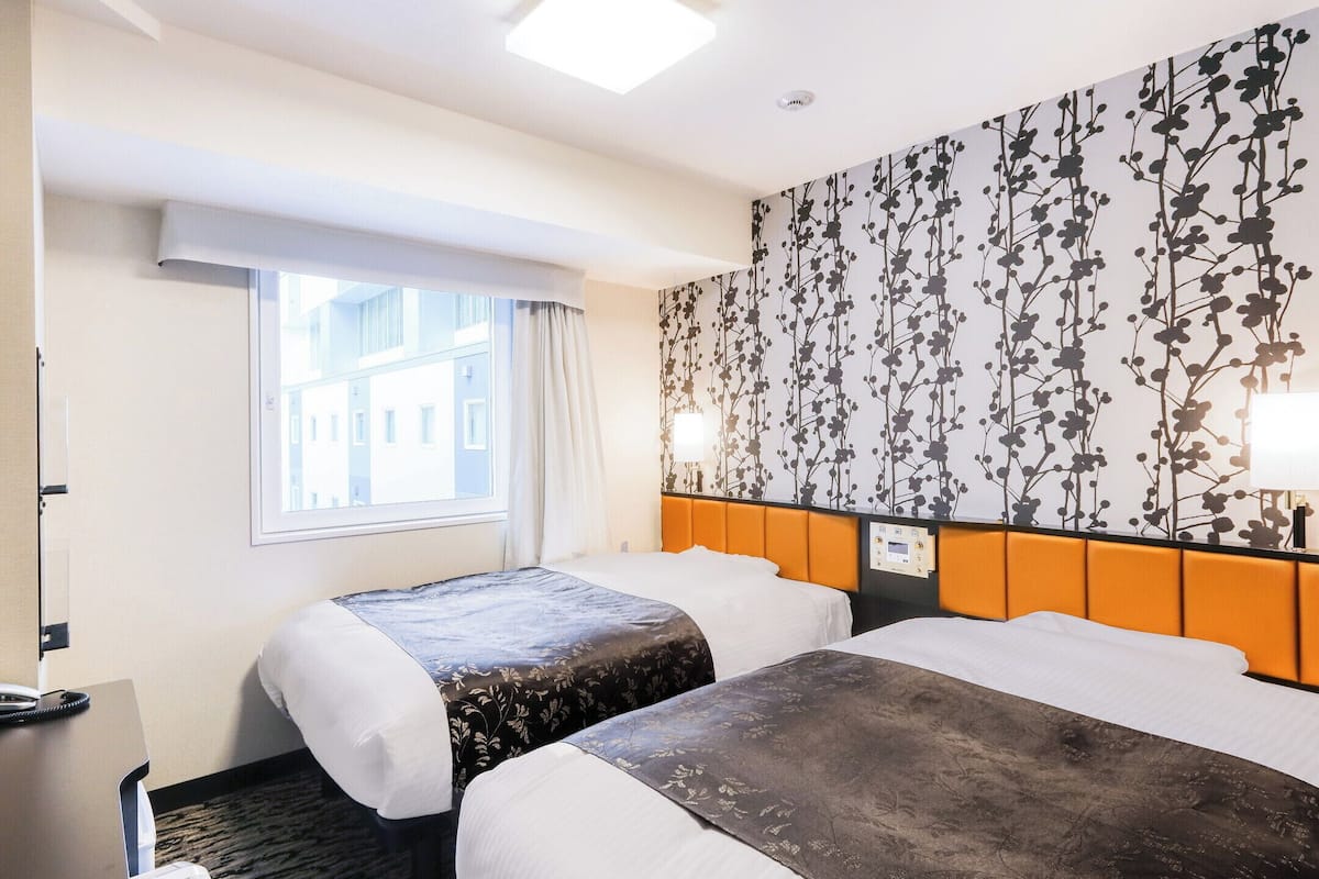 A hotel room with two beds, a TV, a desk with a phone, and a window with curtains.
