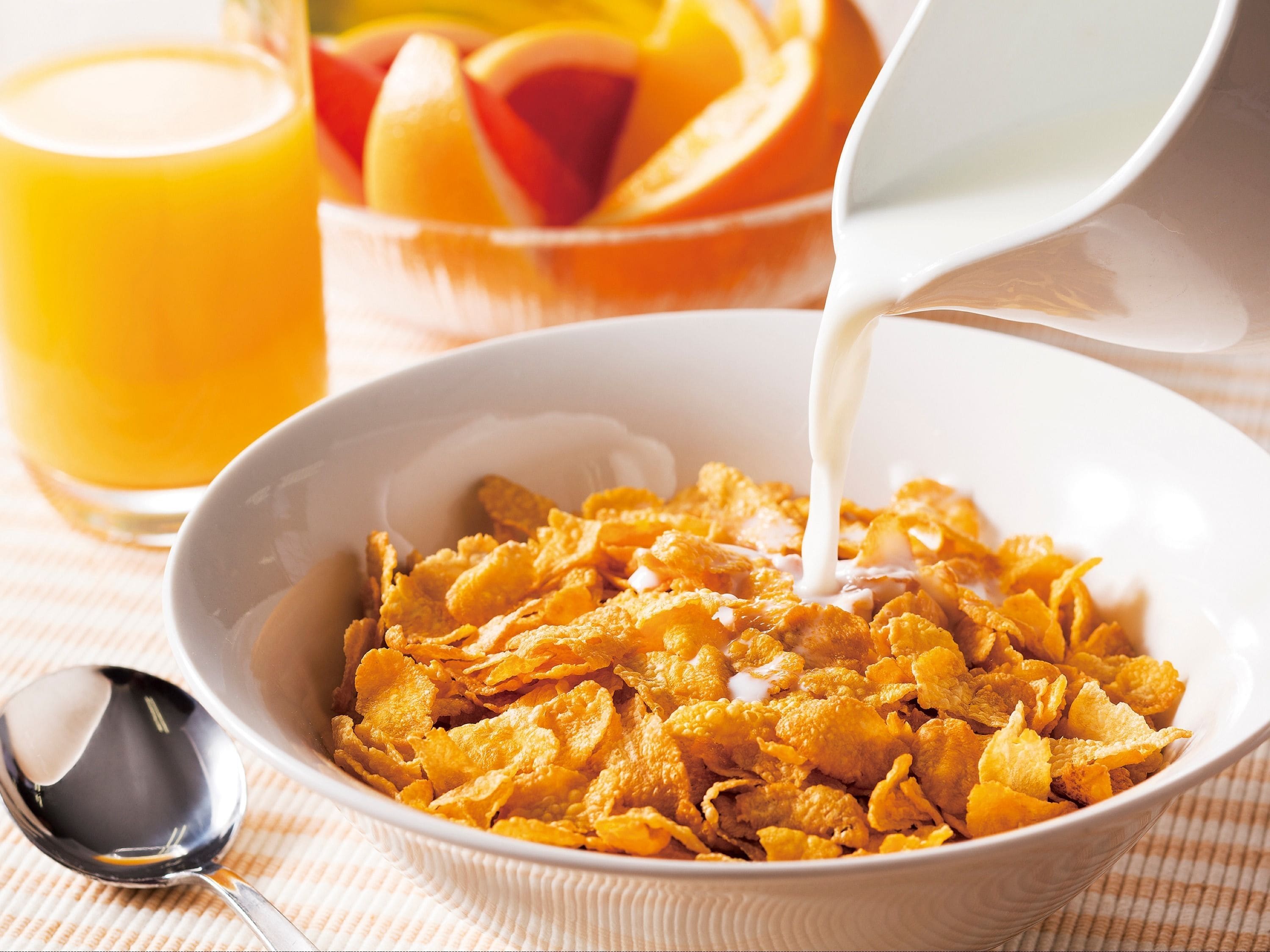 A bowl of cornflakes with milk being poured into it, a glass of orange juice, and a bowl of sliced fruit in the background.