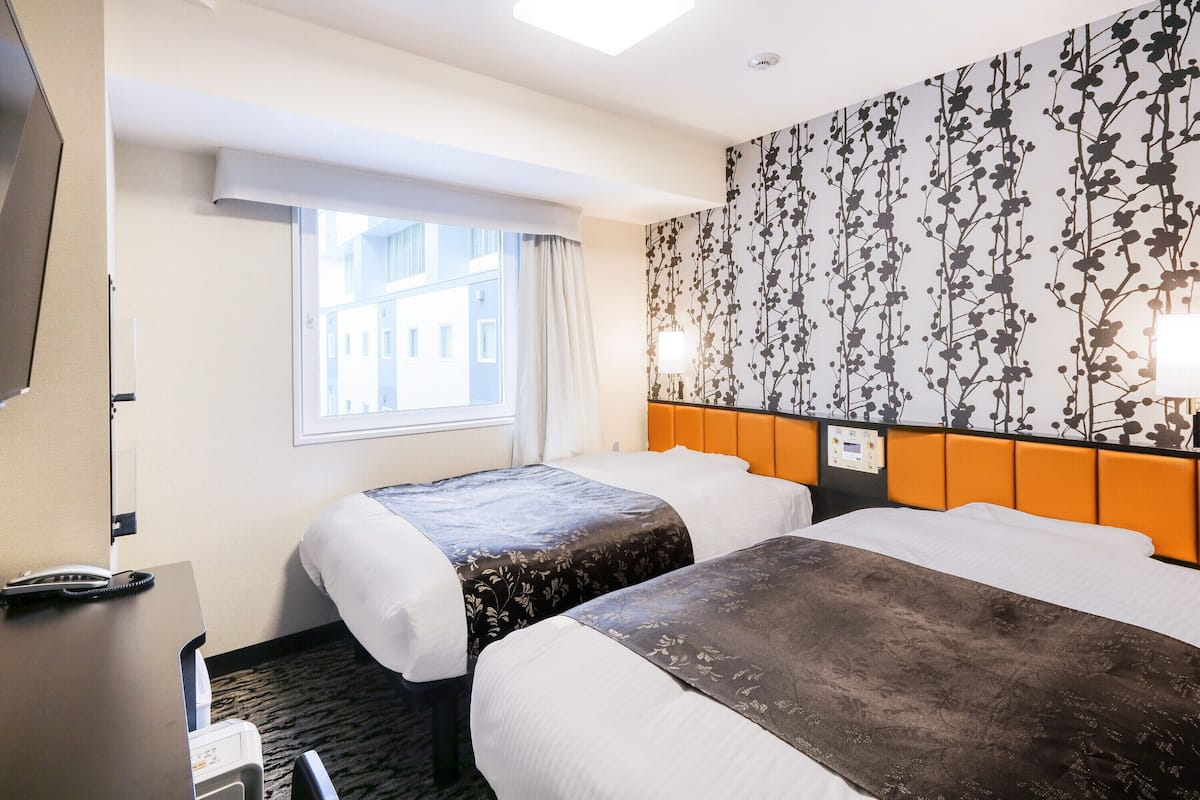Ein Hotelzimmer mit zwei Betten, einem Schreibtisch mit Telefon, einem Fenster mit Blick auf Gebäude und einer Wand mit Blumenmuster.
