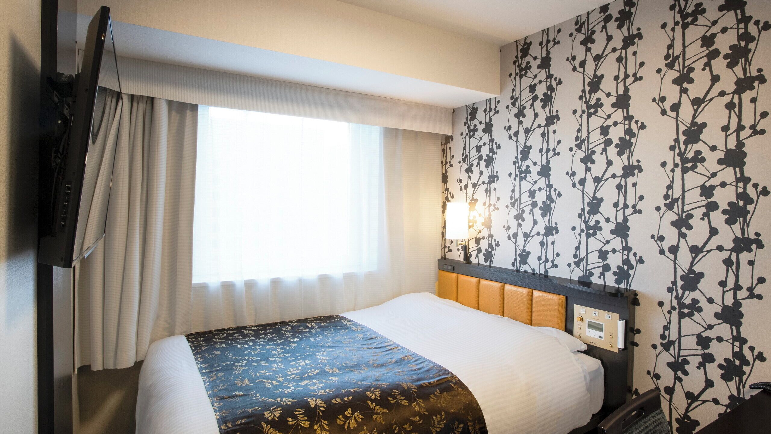 Quarto de hotel com cama, televisão, janela com cortinas e parede com padrão floral.