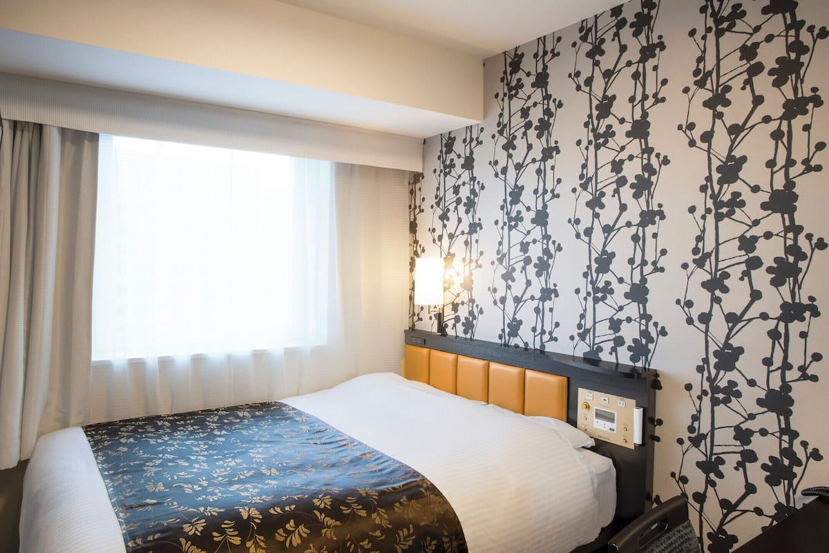 Ein Hotelzimmer mit Bett, Fernseher, einem Fenster mit Vorhängen und einer Wand mit floralem Muster.