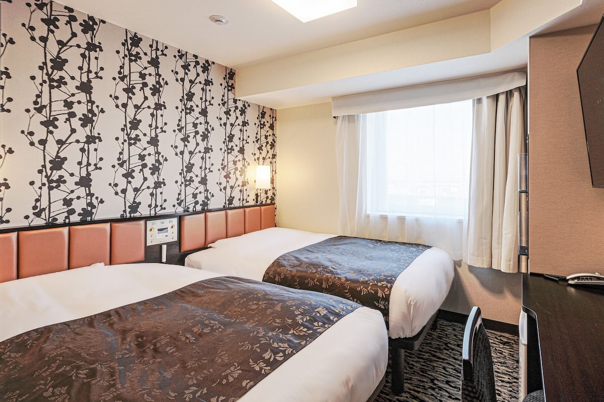 Ein Hotelzimmer mit zwei Betten, einem Kopfteil, einem Nachttisch, einer Lampe, einem Fenster mit Vorhängen und einem Schreibtisch mit Telefon.