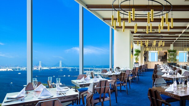 A restaurant with tables set for dining, large windows offering a view of the sea and a bridge, and modern lighting fixtures. - APA Hotel & Resort Yokohama Bay Tower (Yokohama)