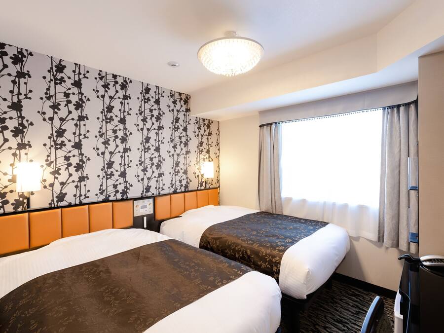 Une chambre d’hôtel avec deux lits, une tête de lit, un panneau de commande fixé au mur, une fenêtre avec des rideaux et un luminaire au plafond.
