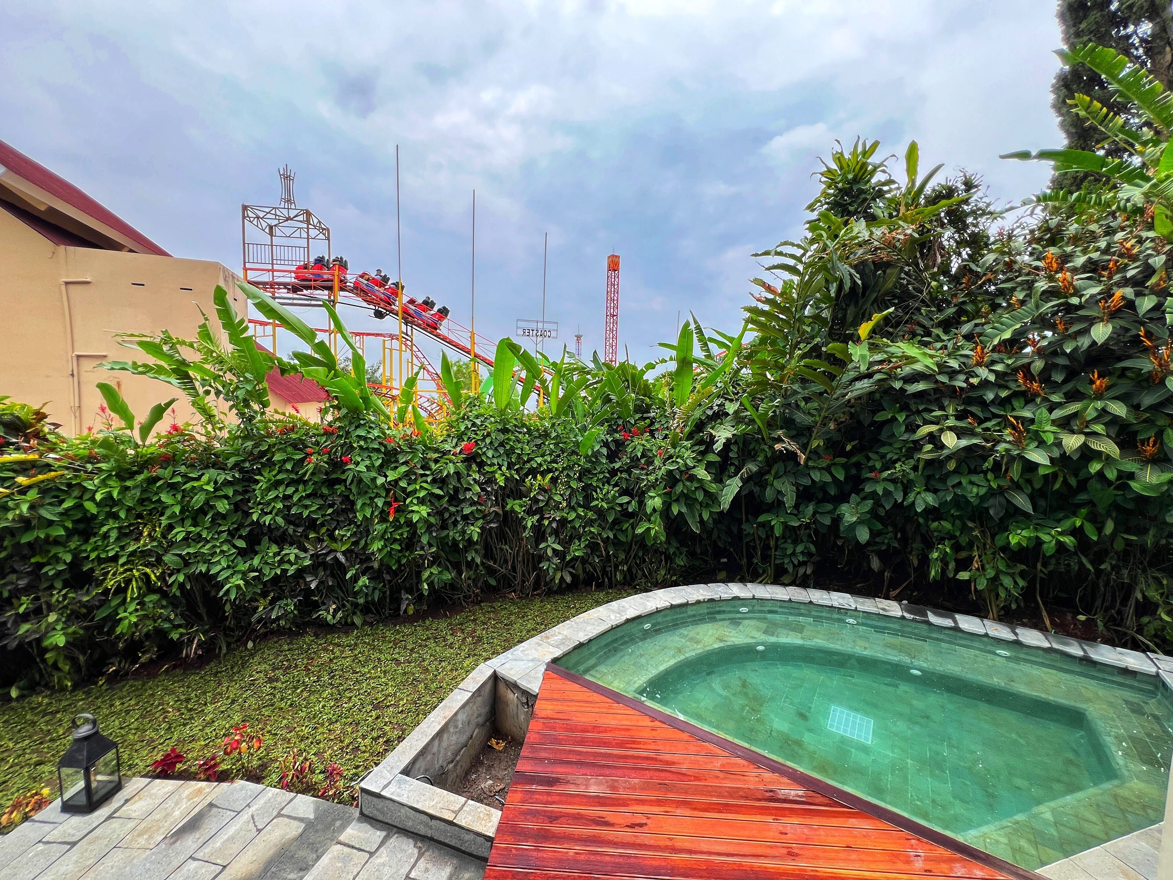 A swimming pool with a wooden deck, surrounded by lush greenery and a roller coaster in the background.