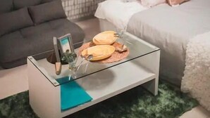 A modern bedroom with a grey sofa, a glass coffee table with decorative items, a bed with white bedding and pink pillows, a green rug, and a brick-patterned wall with a TV mounted above. - Hatch NAGAHORI 202 (Osaka)