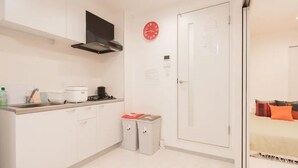 A compact kitchen with white cabinets, a stove, and a microwave. There is a small trash bin and a red clock on the wall. - Hatch NAGAHORI 202 (Osaka)