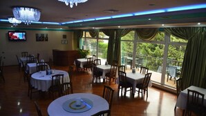 Un restaurant amb taules rodones, caires, un lampadari, un televisor a la pared i grans finestres amb cortines.