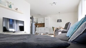 Un soggiorno moderno con una TV a schermo piatto, un divano con cuscini grigi, una zona pranzo con tavolo e sedie, e una cucina sullo sfondo.