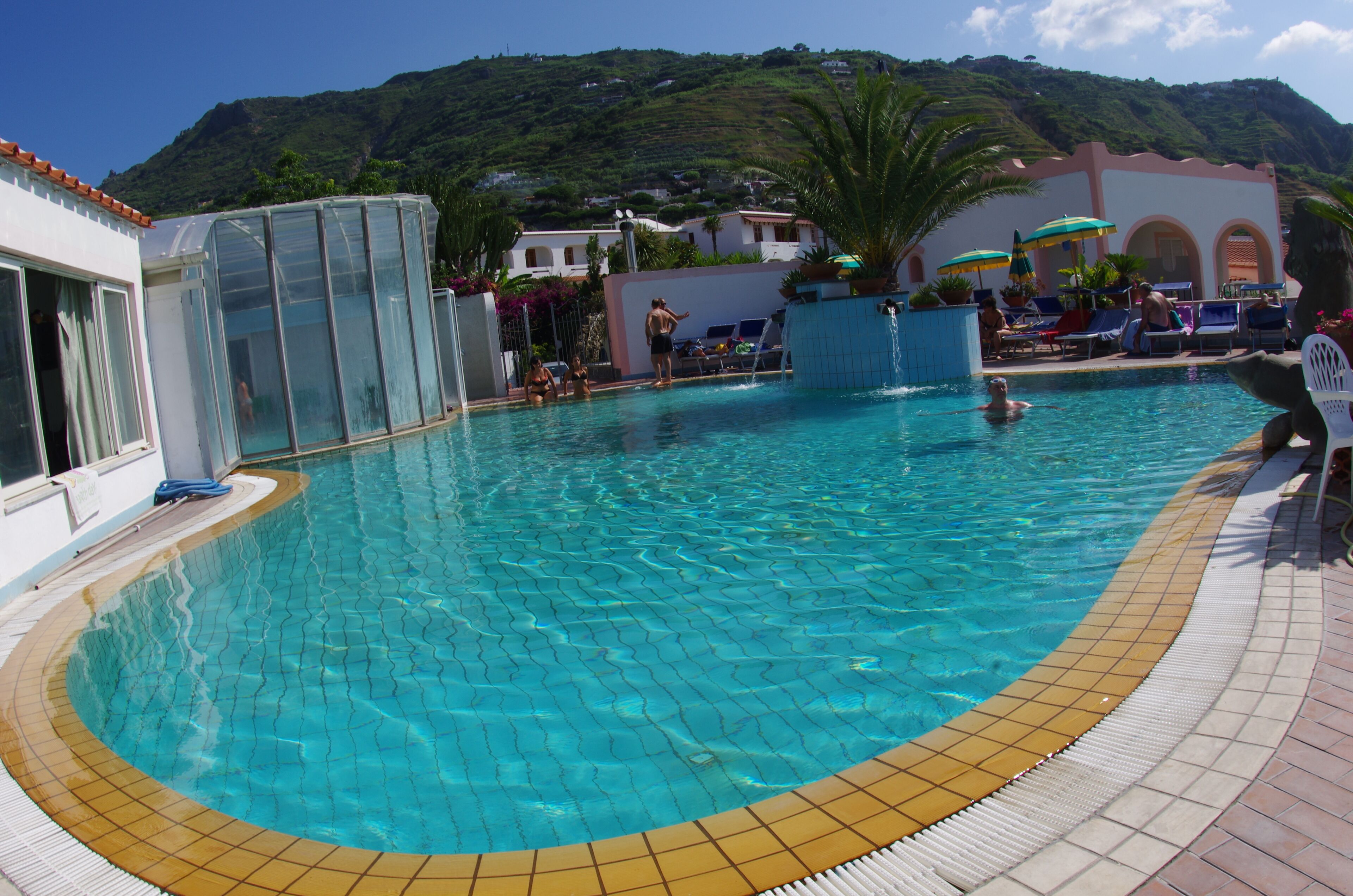 A swimming pool with clear blue water, a tiled deck, and a view of a hillside in the background.