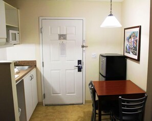 A compact kitchen with a microwave, a refrigerator, a dining table with chairs, a hanging light fixture, and a framed picture on the wall. - Suburban Extended Stay Hotel (Charlotte)