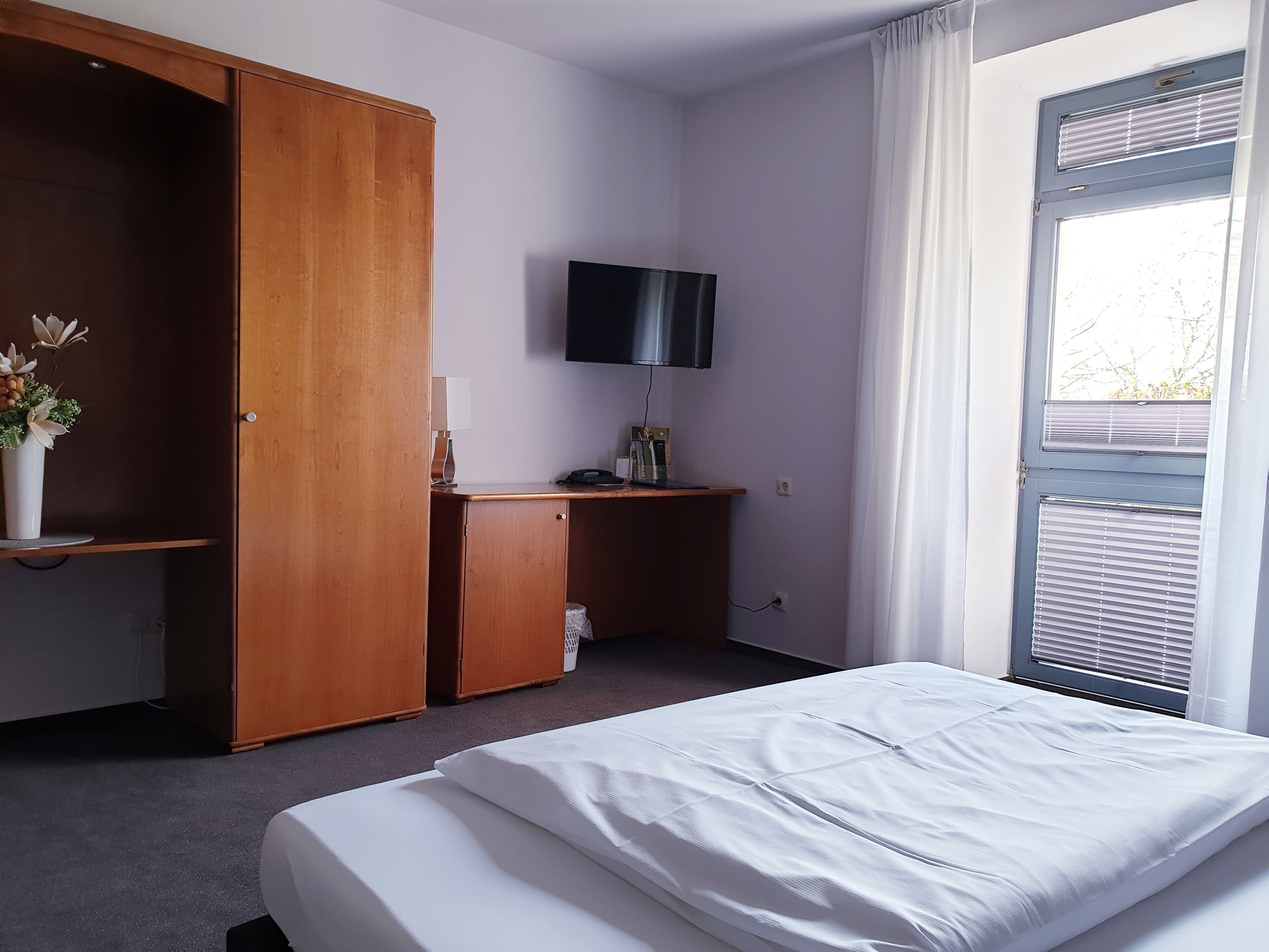 A hotel room with a bed, a wooden wardrobe, a desk with a flat-screen TV, and a window with blinds.