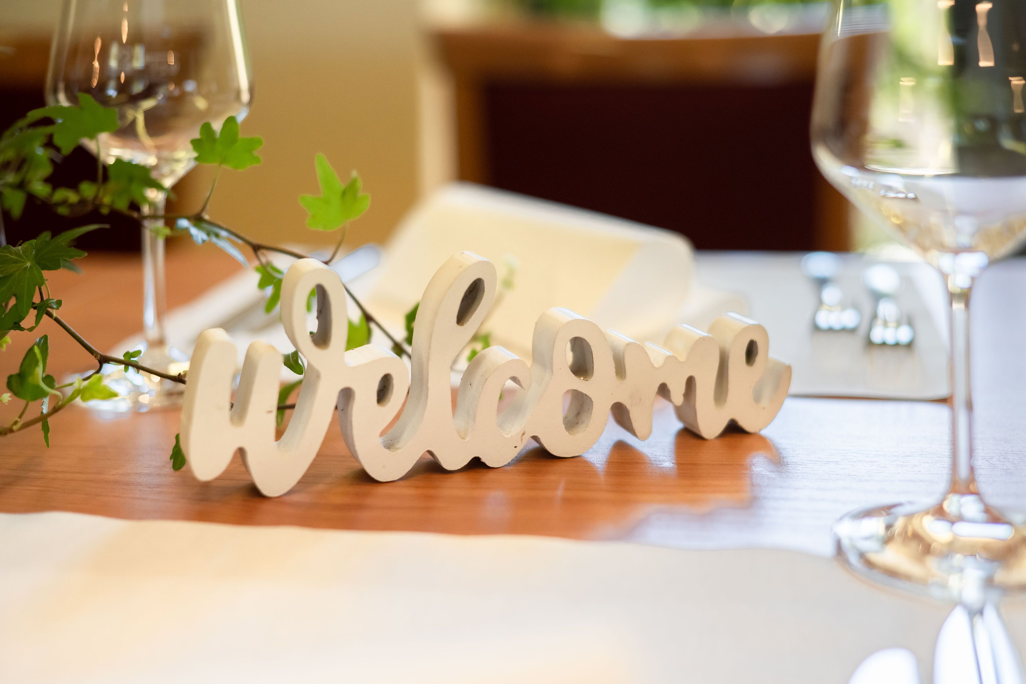 Une table en bois sur laquelle est posée une pancarte blanche avec le mot « Bienvenue », un verre de vin blanc et un verre d’eau.