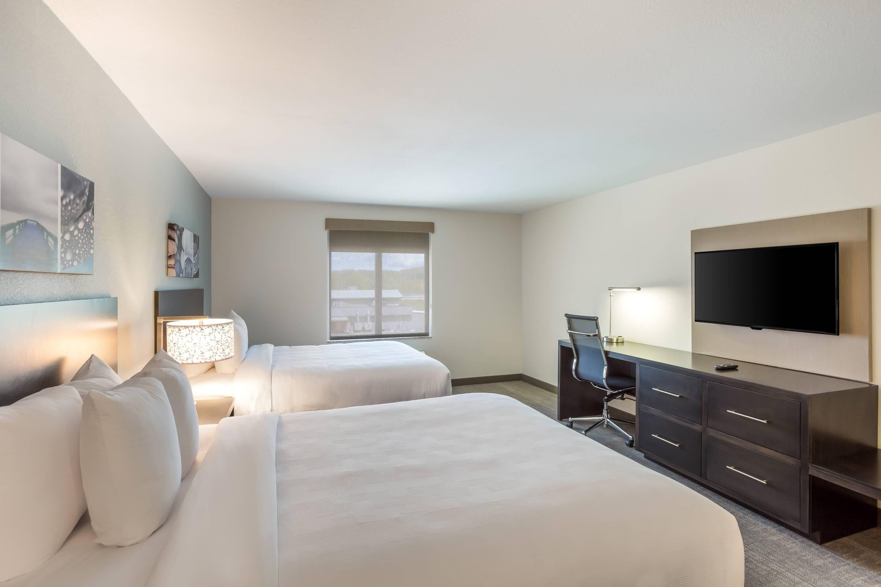 A hotel room with two beds, a desk, a chair, a TV, and artwork on the walls.