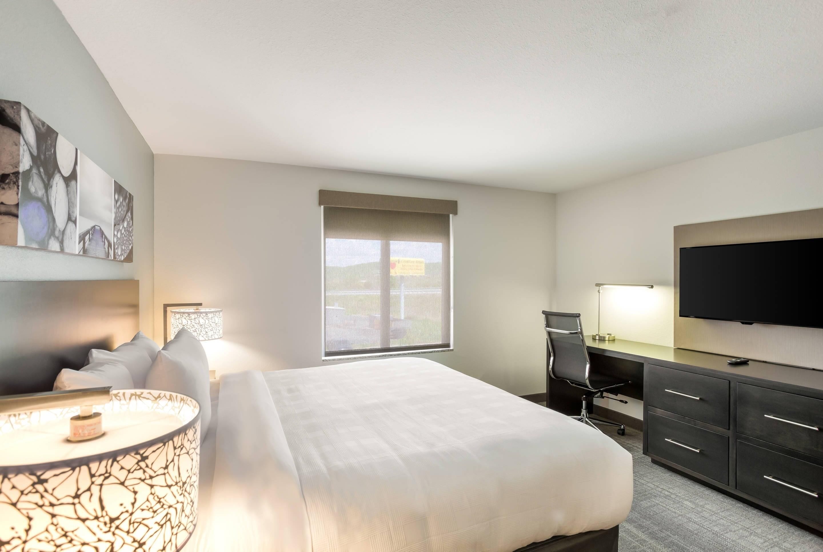 A hotel room with a bed, desk, chair, TV, and artwork on the wall.