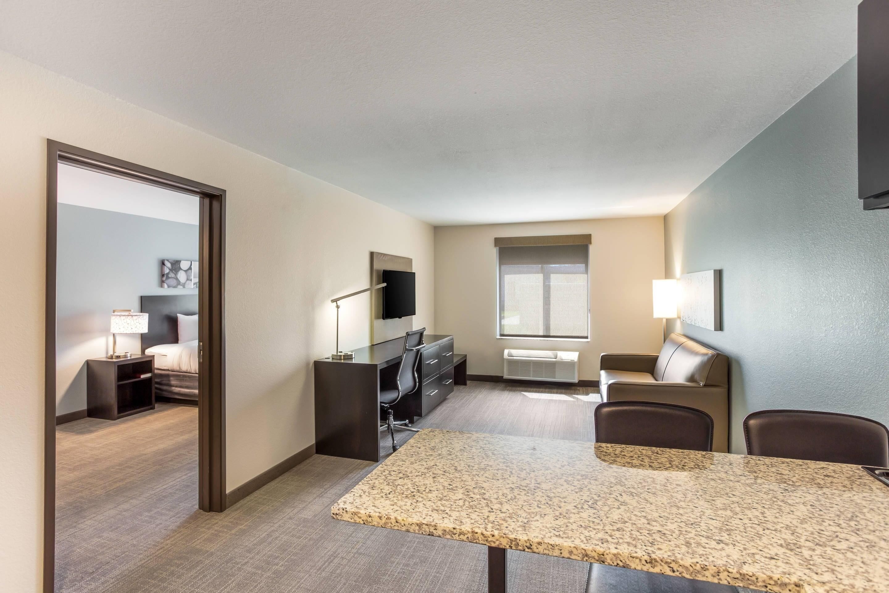 Ein modernes Hotelzimmer mit Essbereich, Fernseher, einer Couch und einem Fenster mit Jalousien.