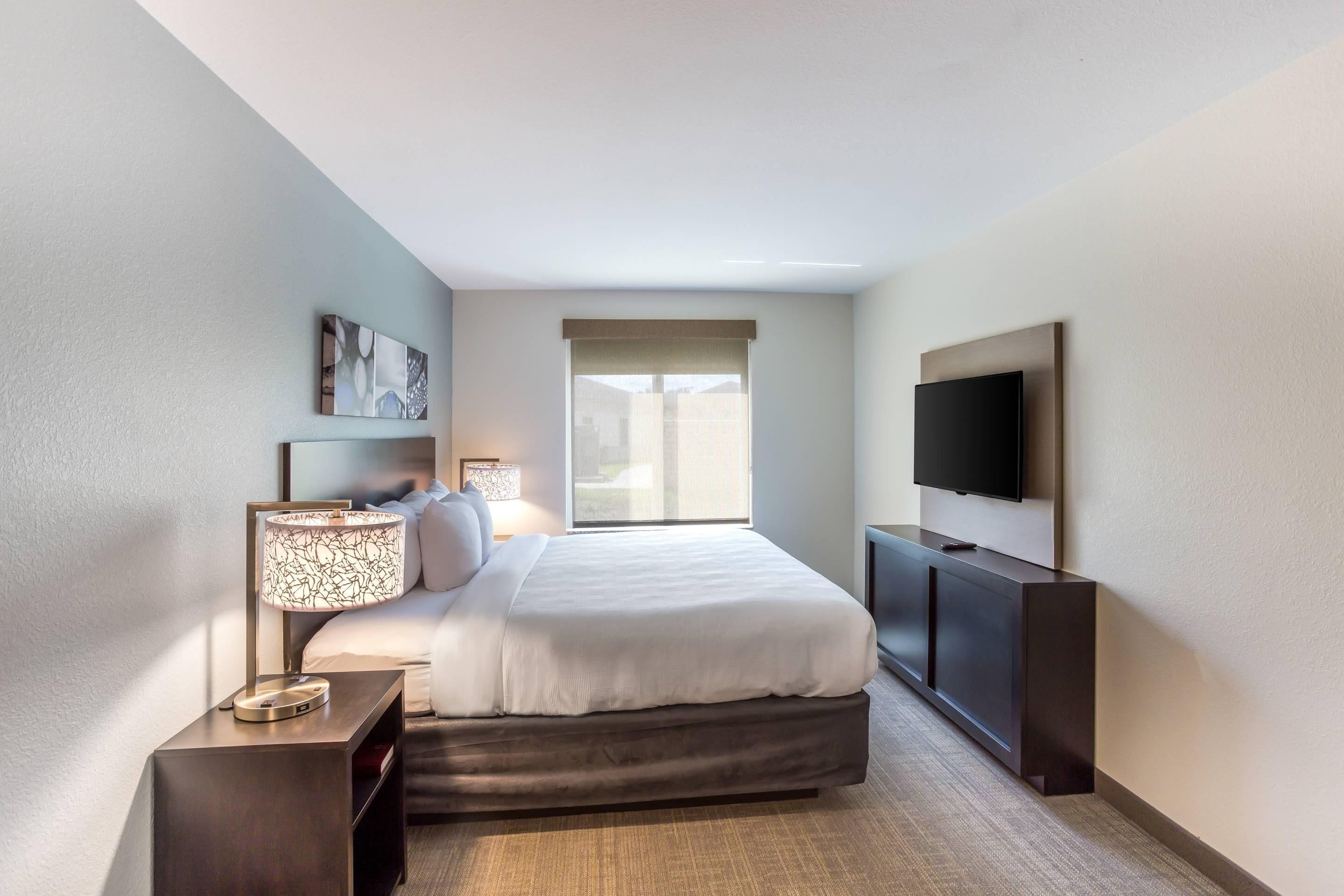 Una habitación de hotel moderna con una cama grande, un televisor montado en la pared y un ventanal que proporciona luz natural.