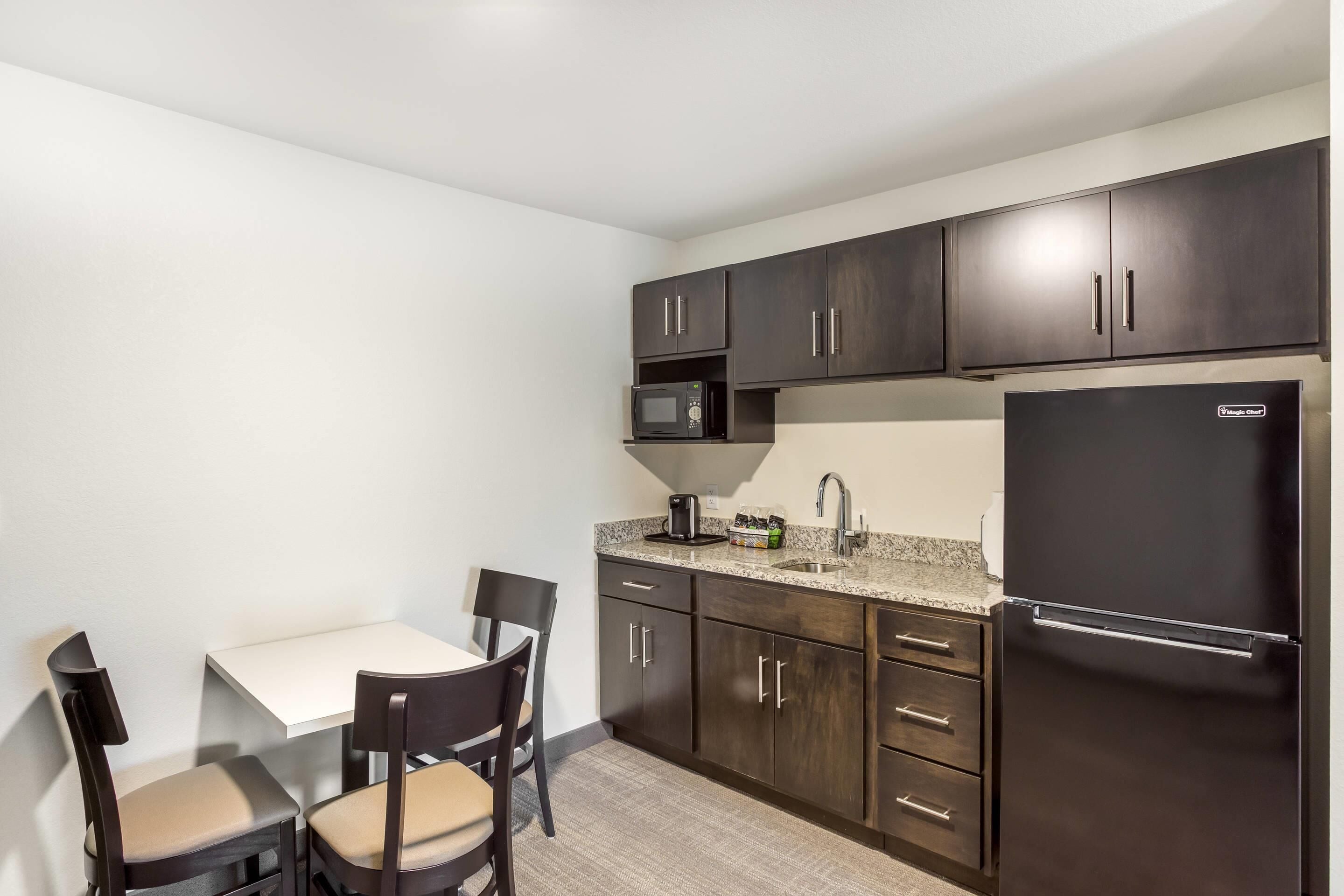 A compact kitchen with dark cabinets, a microwave, a refrigerator, and a small dining area with two chairs and a table.