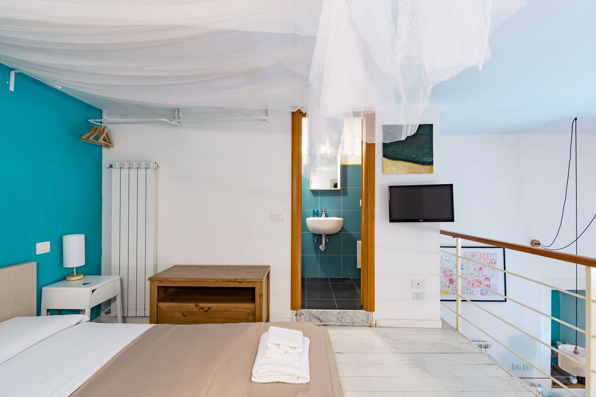 Une chambre à coucher avec un lit, une table de chevet et un banc en bois. On aperçoit une salle de bain à travers une porte ouverte, avec un lavabo et un miroir.