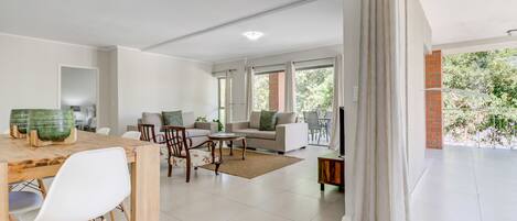 A spacious living area with a dining table, sofa, and a balcony with a view of greenery.