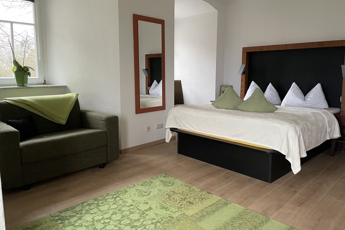 A bedroom with a bed, a green armchair, a large mirror, and a window with a plant.