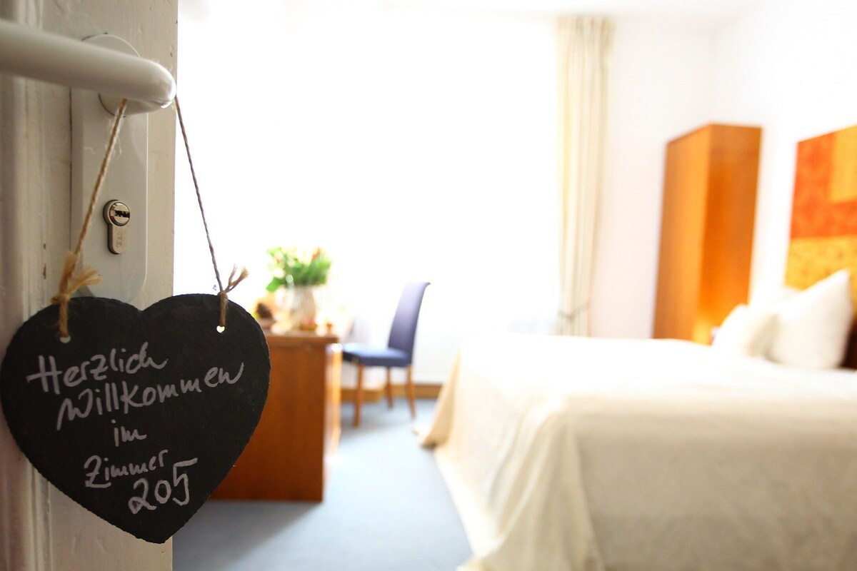 A hotel room with a bed, a chair, a small table with a plant, and a heart-shaped welcome sign.