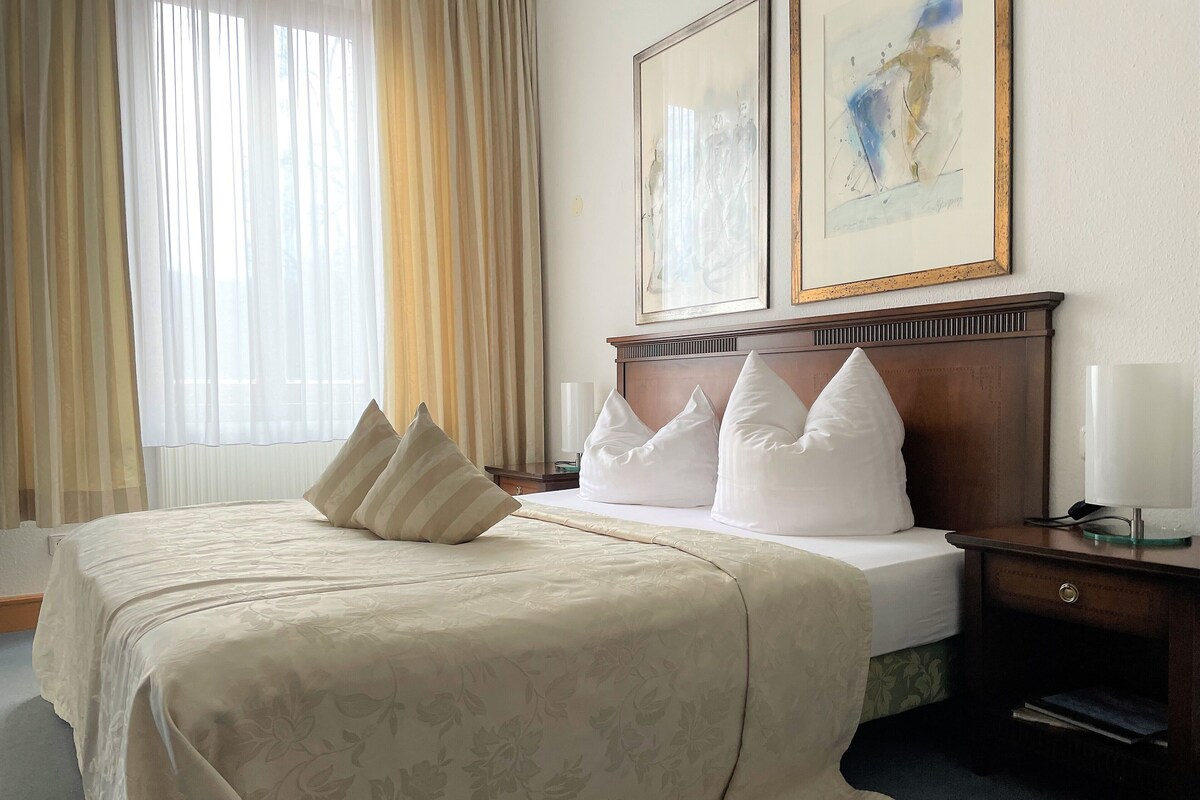 A hotel room with a neatly made bed, a nightstand with a lamp, and two framed artworks on the wall.