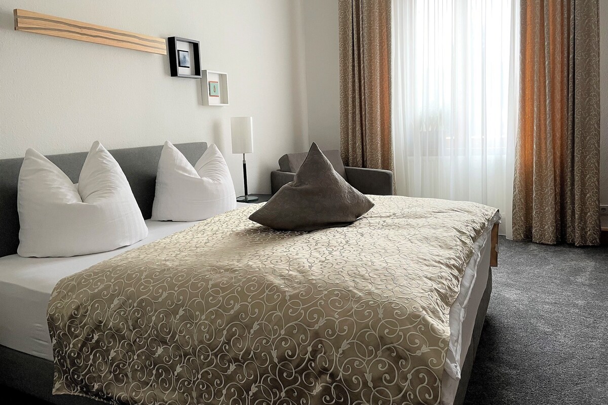 A hotel room with a bed, two pillows, a bedside table with a lamp, a picture frame, and a window with curtains.