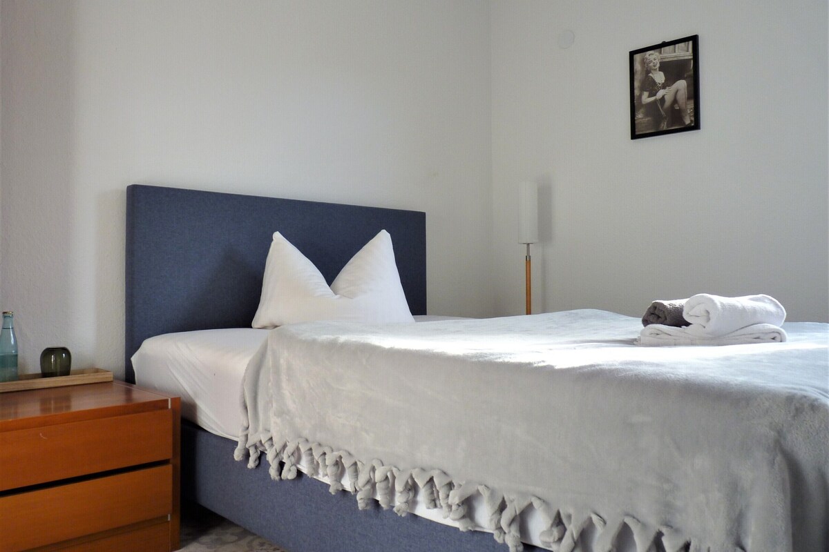 A neatly made bed with white linens and a blue headboard, a wooden nightstand with a bottle and a small container, and a framed picture on the wall.