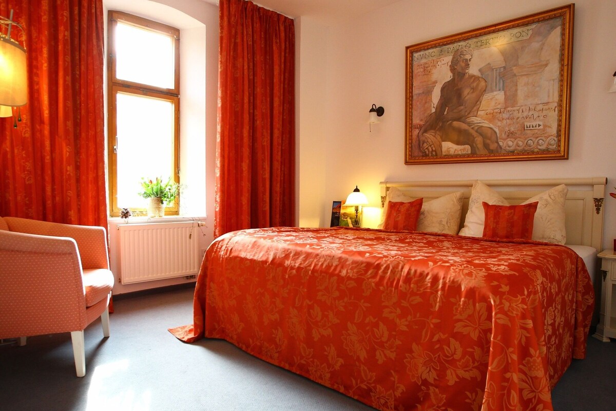 A hotel room with a large bed, a chair, a window with curtains, a painting on the wall, and a lamp on the nightstand.