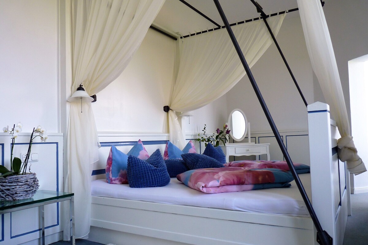 A bedroom with a canopy bed, bedside table, lamp, mirror, and a small potted plant.