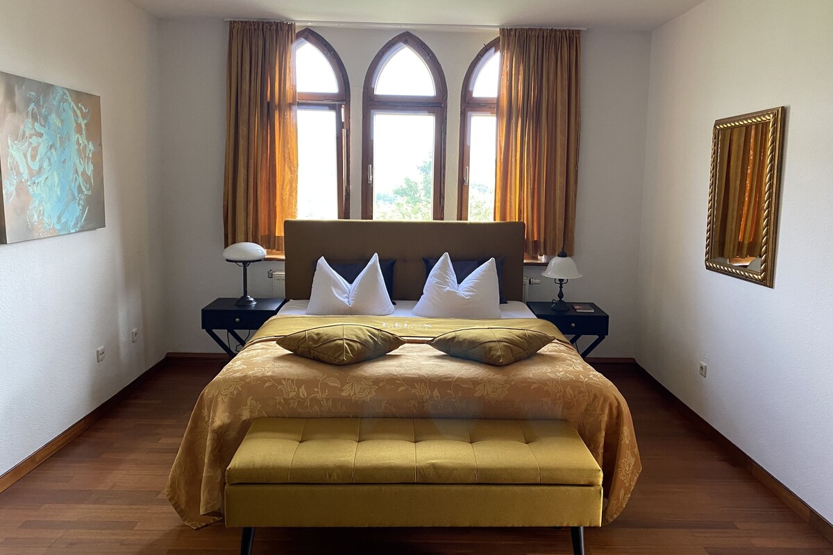 A bedroom with a large bed, two bedside tables, a bench, and three windows with curtains.