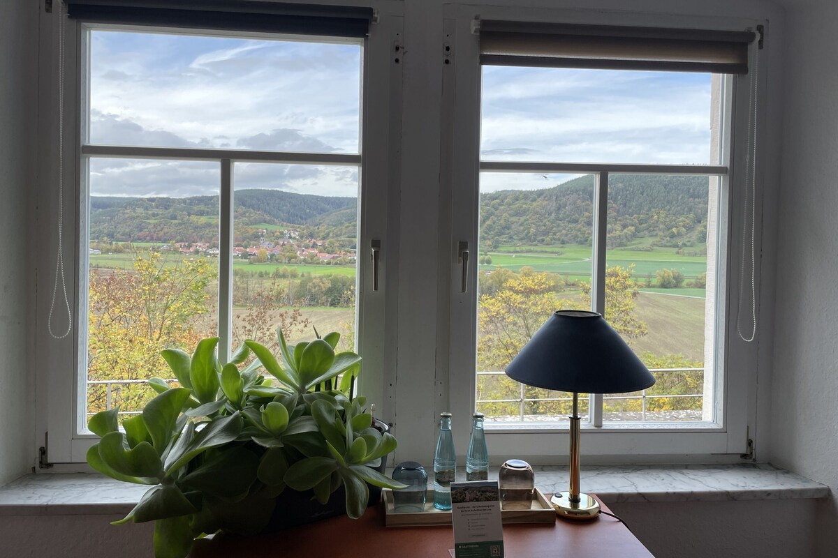 A room with two large windows offering a view of a green landscape with hills and trees.