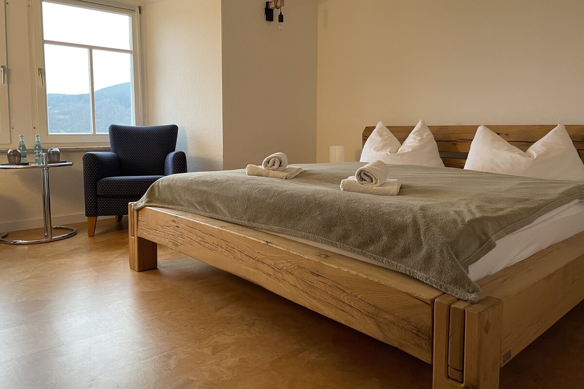 A neatly made bed with a wooden headboard, a chair with a cushion, and a small table with bottles and glasses.