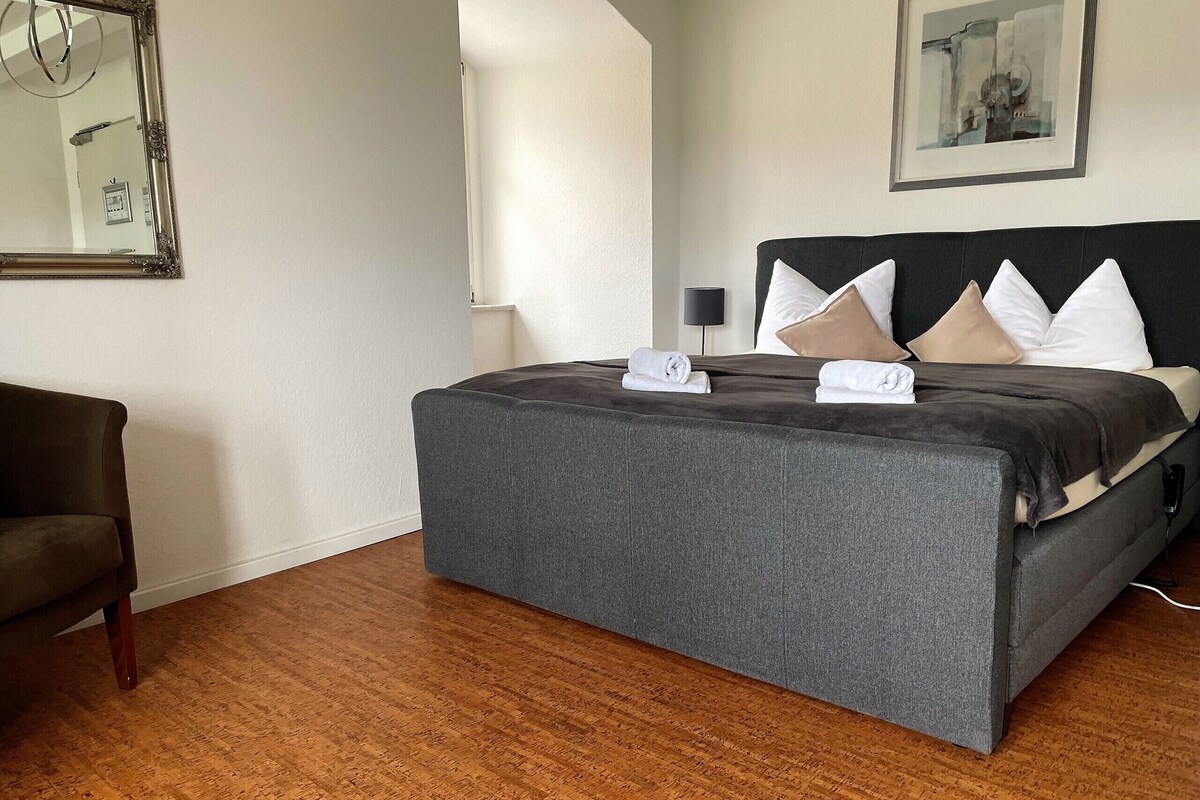 A neatly made bed with a dark headboard, a grey upholstered bench, and a wooden floor.